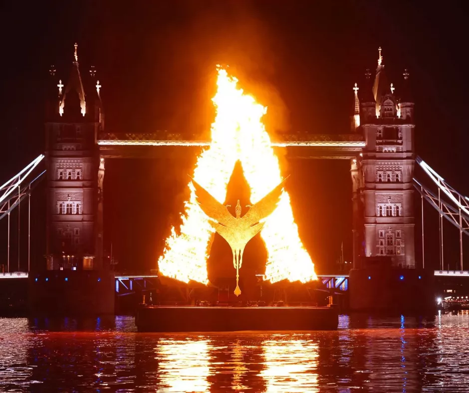 A large fiery sculpture shaped like a glowing letter “A” burns brightly on a floating platform on the River Thames at night. At the center of the flame is a stylized bird figure with wings spread. Tower Bridge is illuminated in the background.